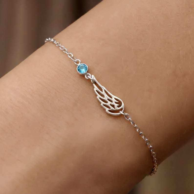 May include: A delicate silver bracelet featuring a small, round, light blue gemstone and a stylized wing charm. The bracelet is made of a fine chain and is designed to be worn on the wrist. The wing charm has intricate cut-out details.