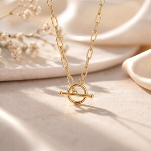 May include: A gold-toned necklace featuring a series of elongated oval links connected to a circular toggle clasp with a horizontal bar. The metal has a polished finish, and the chain design is uniform throughout the visible length of the piece.