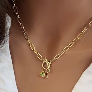 May include: A gold chain necklace with a toggle clasp and a charm featuring a green gemstone and the letter J.
