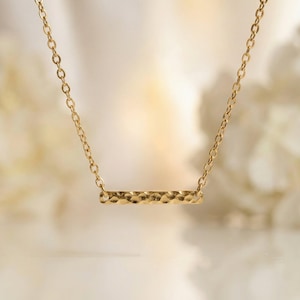 Puede incluir: Un collar de tono dorado que presenta un colgante de barra horizontal con un acabado texturizado y martillado. El colgante est&aacute; suspendido de una cadena de eslabones fina y delicada que se conecta a cada extremo de la barra, creando una joya simple.