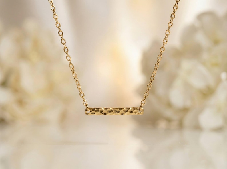 Puede incluir: Un collar de tono dorado que presenta un colgante de barra horizontal con un acabado texturizado y martillado. El colgante est&aacute; suspendido de una cadena de eslabones fina y delicada que se conecta a cada extremo de la barra, creando una joya simple.