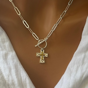 May include: Silver-toned chain necklace with a toggle clasp and a cross pendant. The cross features a textured silver frame with a gold-colored center. The necklace is displayed against a white fabric background.