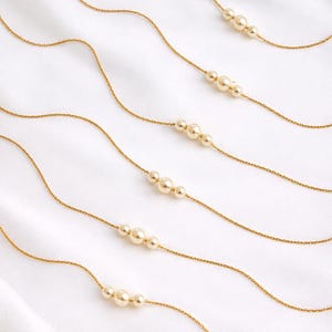 May include: Delicate gold necklaces with varying arrangements of cream-colored pearls. The necklaces are displayed on a white, textured surface, creating a minimalist and elegant aesthetic. The fine gold chains add a touch of sophistication.