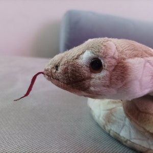 May include: A plush snake toy with a brown and pink patterned body and a red tongue. The snake is curled up on a light gray surface.