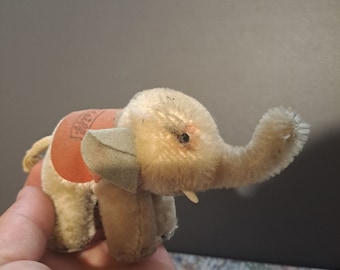Pode incluir: Um pequeno elefante de pelúcia bege com uma sela marrom e uma tromba verde claro. O elefante tem um nariz rosa e uma única presa branca.