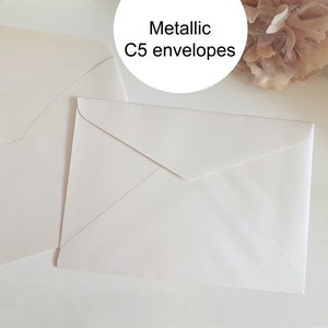 May include: Two white C5 envelopes with a metallic finish. The envelopes are lying flat on a white surface.