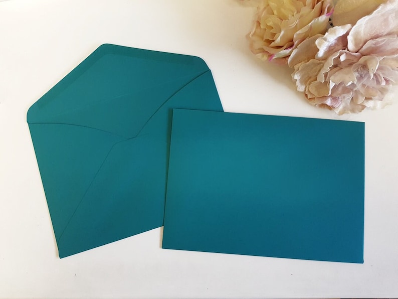 May include: A teal stationery set comprising an envelope and a blank card. The envelope is open, displaying its interior. The rectangular card is ready for a handwritten message. Soft pink and cream-coloured flowers are in the background.