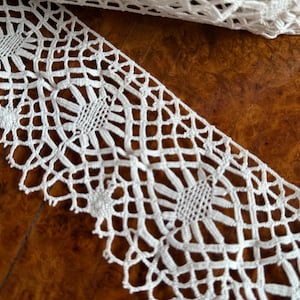 Puede incluir: Borde de encaje blanco tejido a crochet con un delicado patrón floral. El encaje tiene un diseño repetitivo de pequeñas flores y una cuadrícula en forma de diamante.