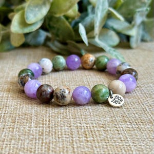 May include: A beaded bracelet featuring a variety of colorful, round gemstones in shades of purple, green, brown, and cream. The bracelet includes a small, silver-colored charm with a lotus flower design. The bracelet is displayed on a neutral, textured surface.