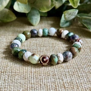 Puede incluir: Una pulsera de cuentas multicolores. La pulsera presenta cuentas verdes, blancas, marrones y estampadas. Las cuentas están ensartadas para formar una forma circular. La pulsera se muestra sobre una superficie texturizada y neutra.