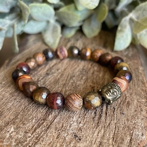 Może przedstawiać: Bransoletka z koralików z brązową głową Buddy i mieszanką brązowych, czerwonych i złotych koralików. Koraliki mają różne faktury, w tym fasetowane, prążkowane i gładkie wykończenia. Bransoletka jest prezentowana na drewnianej powierzchni.