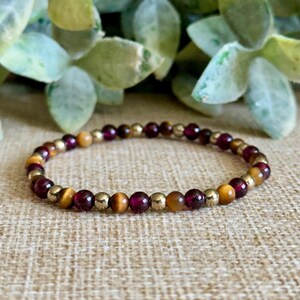 May include: A beaded bracelet featuring a mix of gold, burgundy, and amber-colored beads. The bracelet is strung on an elastic band and is shown against a neutral background with green foliage in the background.