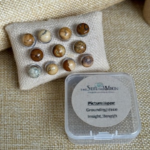 May include: A set of 10 picture jasper push pins in a clear plastic container. The push pins are brown, tan, and white with black markings. The container has a label that reads "The Sun and the Moon" and "Picture Jasper Grounding, Peace Insight, Strength".