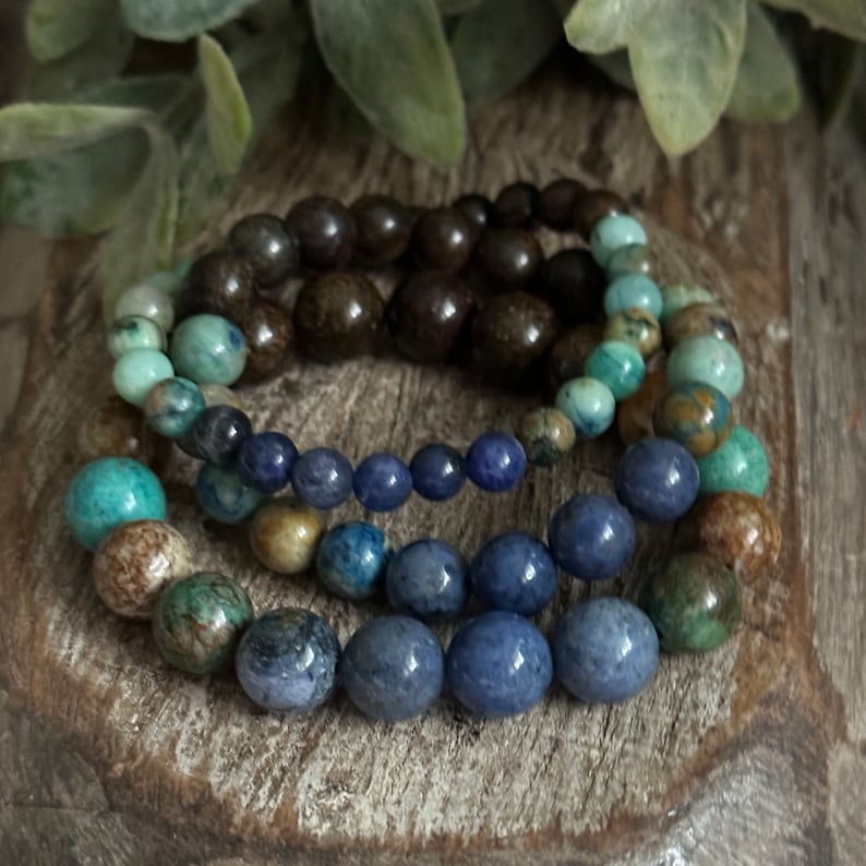 May include: Four stacked bracelets made of blue, green, and brown beads. The bracelets are made of different materials, including wood and stone.
