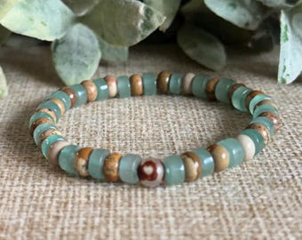 Puede incluir: Un brazalete de cuentas con cuentas alternas de color verde claro, marrón y blanquecino. Las cuentas son pequeñas y redondas, ensartadas en una banda elástica. El brazalete se muestra sobre un fondo neutro. Circunferencia de unos 18 cm.
