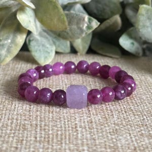 May include: A purple beaded bracelet with a light purple square-shaped center bead. The round beads are a deep purple color. The bracelet is displayed on a textured, neutral-colored surface.