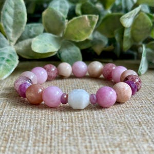 May include: A beaded bracelet featuring a variety of pink, peach, and white gemstones. The bracelet includes round and faceted beads in shades of pink, peach, and white, strung together on an elastic band. The bracelet is displayed on a textured surface.