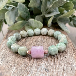 Könnte beinhalten: Ein Armband mit hellgrünen, facettierten Perlen und einer zentralen, hellvioletten, rechteckigen Perle. Das Armband ist auf einem Gummiband aufgezogen. Das Armband wird auf einer Holzoberfläche präsentiert.