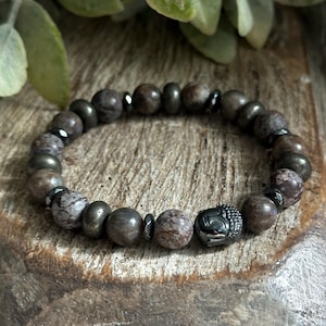 Puede incluir: Un brazalete de cuentas marrones y grises con un dije de cabeza de Buda negro. Las cuentas están hechas de piedra natural y el dije está hecho de metal.