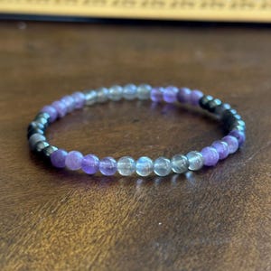 May include: A beaded bracelet featuring a mix of purple, gray, and black beads. The bracelet is made of small, round beads strung together to form a flexible band. The beads have a polished, shiny appearance.