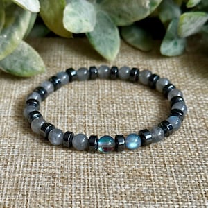 Può includere: Un braccialetto di perline con perline grigie e nere. Il braccialetto ha un mix di perline grigie rotonde e perline cilindriche nere. Alcune perline iridescenti aggiungono un tocco di colore. Il braccialetto è esposto su una superficie dai toni neutri.