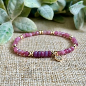 May include: A pink gemstone bracelet with gold accents and a heart charm. The bracelet features small, round pink beads interspersed with gold-colored beads. A cluster of darker pink beads is in the center. A small gold heart charm is attached.