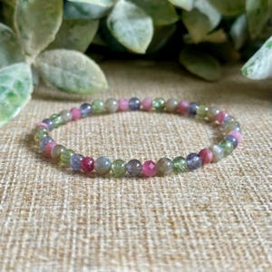 May include: A beaded bracelet featuring a variety of colorful gemstones. The bracelet includes pink, green, blue, and gray beads. The bracelet is displayed on a textured, tan surface, with green foliage in the background.