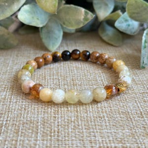 Op de afbeelding: Een meerkleurige kralen armband met een gouden bedel. De armband heeft een verloop van bruine, groene en witte kralen.