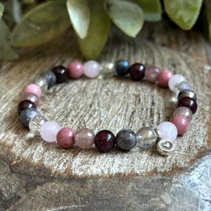 Puede incluir: Un brazalete hecho con cuentas de piedras preciosas de color rosa, rojo y gris. El brazalete tiene un dije de plata con forma de corazón.
