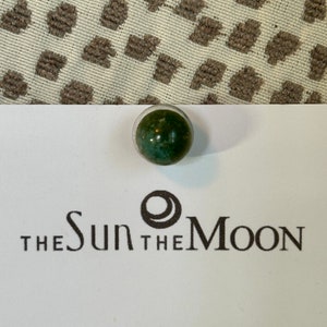 May include: A green and white marble with a dark green swirl pattern. The marble is sitting on a white card with the text "THE SUN THE MOON" printed on it.