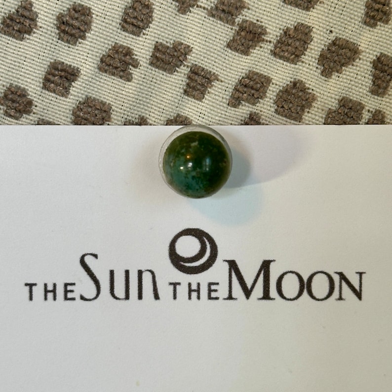 May include: A green and white marble with a dark green swirl pattern. The marble is sitting on a white card with the text "THE SUN THE MOON" printed on it.