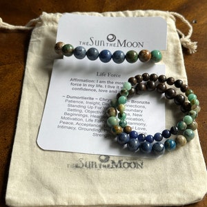 May include: Three beaded bracelets with different colored stones. The bracelets are stacked on top of each other. The top bracelet has green and brown stones, the middle bracelet has brown stones, and the bottom bracelet has blue and green stones. The bracelets are on a white background with text that reads "The Sun The Moon" and "Life Force".
