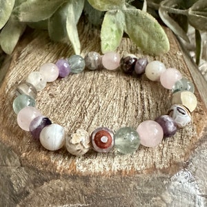 Op de afbeelding: Een armband gemaakt van verschillende halfedelstenen, waaronder roze, groene, witte en paarse kralen. De armband is gemaakt van natuurlijke stenen en heeft een unieke, aardse uitstraling.