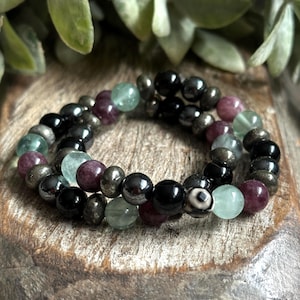 Puede incluir: Un brazalete de múltiples hilos con cuentas negras, verdes y moradas. El brazalete está hecho de varias piedras preciosas, incluyendo hematita, pirita y fluorita.