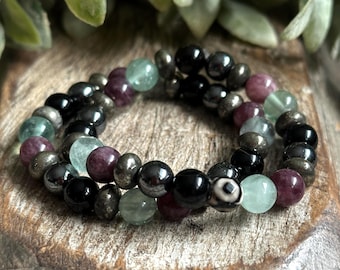 May include: A multi-strand bracelet with black, green, and purple beads. The bracelet is made of various gemstones, including hematite, pyrite, and fluorite.