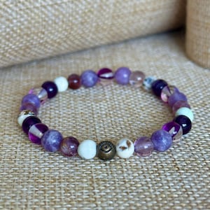 May include: A purple and white beaded bracelet with a gold charm featuring an eye design. The bracelet is made with various types of stones, including amethyst, agate, and quartz.