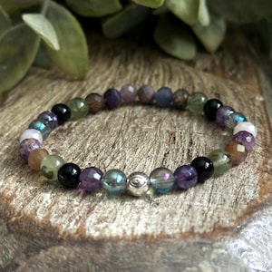 May include: A multi-colored beaded bracelet with a silver charm. The beads are various shades of purple, green, brown, and black. The bracelet is on a wooden surface.
