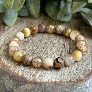 Puede incluir: Un brazalete de piedras preciosas marrón, beige y dorado con una variedad de patrones. El brazalete está hecho de cuentas redondas y está enhebrado en un cordón elástico transparente.