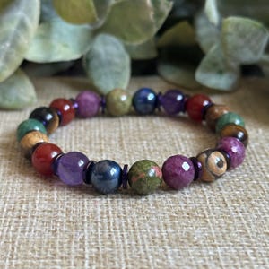 May include: A beaded bracelet featuring a variety of colorful gemstones. The bracelet includes beads in shades of red, purple, green, blue, and brown. The beads are strung together with a purple elastic cord. The bracelet is displayed on a tan surface.