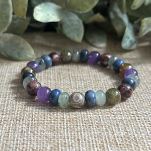 May include: A beaded bracelet featuring a variety of colorful gemstones. The bracelet includes purple, blue, green, and brown beads. The focal bead has a unique pattern. The bracelet is displayed on a neutral-toned surface.