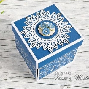 May include: A blue square box with a white lace design and a floral embellishment on the top. The box is decorated with a white and blue pattern and the text "designed by Fame's World".