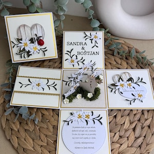 May include: A white card with a pop-up design featuring a couple standing in front of a wooden heart. The card is decorated with white flowers and green leaves. The text on the card reads "Sandra & Boštjan".