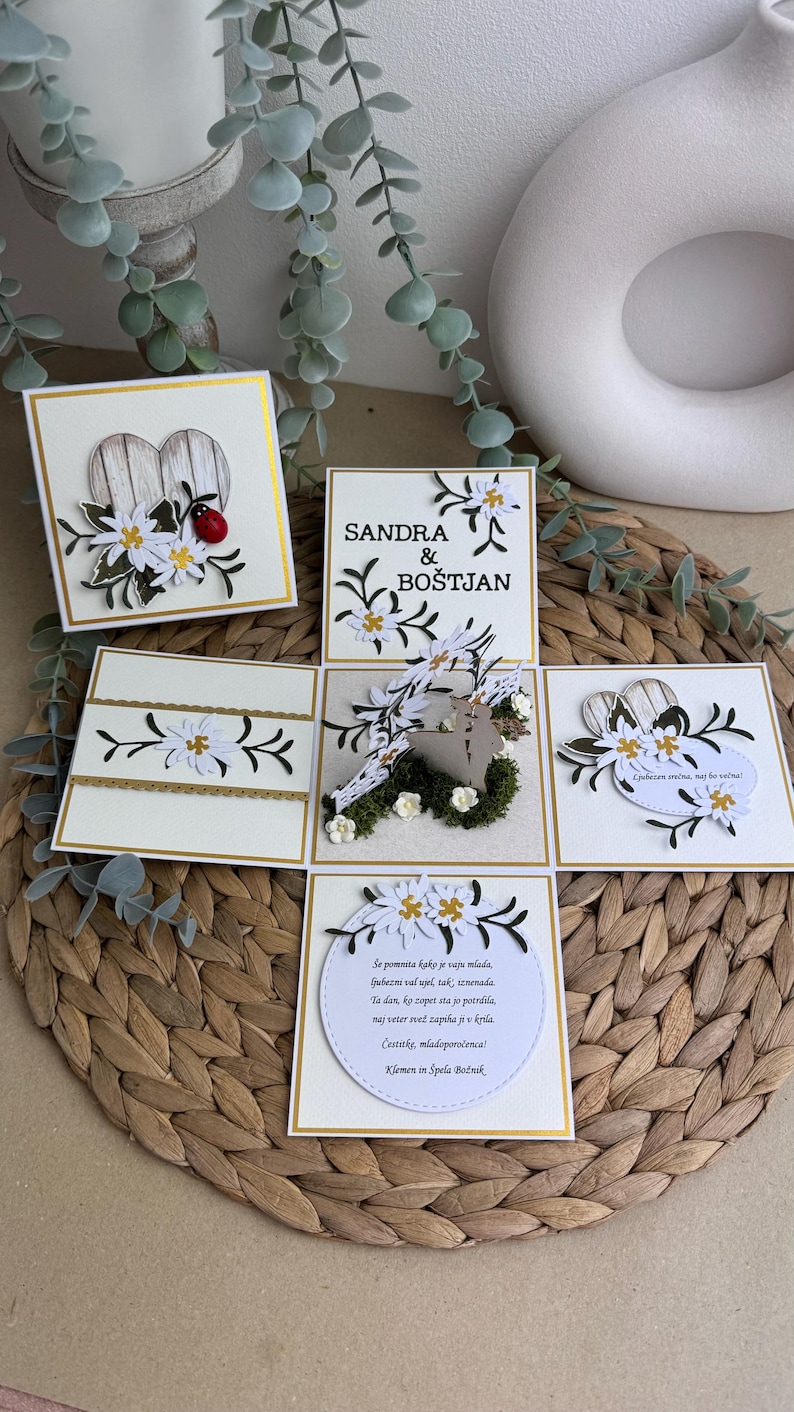 Op de afbeelding: Een witte kaart met een pop-up ontwerp met een stel dat voor een houten hart staat. De kaart is versierd met witte bloemen en groene bladeren. De tekst op de kaart luidt "Sandra & Bo&scaron;tjan".
