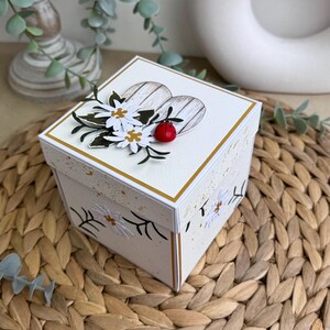 Op de afbeelding: Een witte vierkante doos met een hartvormig ontwerp aan de bovenkant. Het hart is gemaakt van hout en heeft een witgekleurde afwerking. De doos is versierd met witte bloemen, groene bladeren en een rode lieveheersbeestje. De doos staat op een geweven strooien mat.