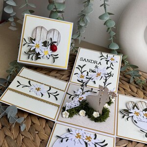 Op de afbeelding: Een witte en gouden trouwkaart met een pop-up ontwerp. De kaart toont een stel dat voor een wit hek met bloemen en groen staat. De kaart bevat ook een wit hart met een lieveheersbeestje en een bericht dat luidt: "Se pomnita kako je vaju mlada, ljubezni val ujel, tak, iznenada. Ta dan, ko zopet sta jo potrdila, naj veter svež zapiha ji v krila. Čestitke, mladoporočenca! Klemen in Spela Božnik"