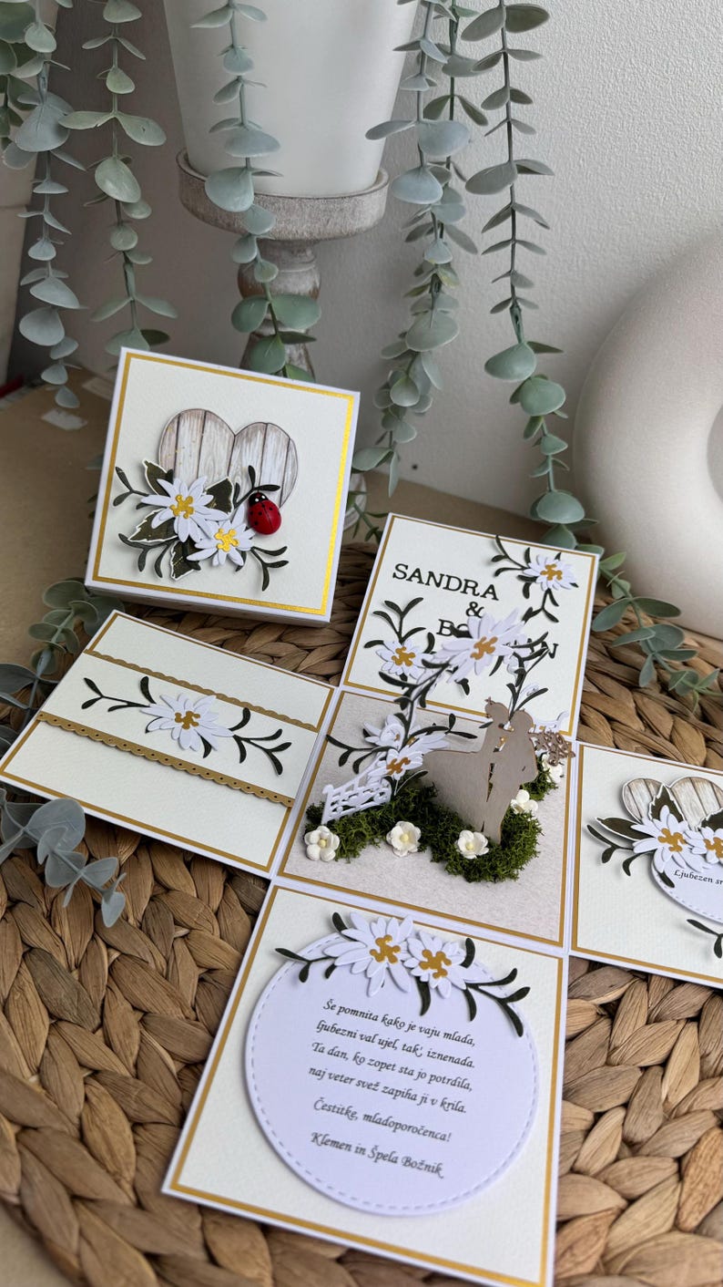 Op de afbeelding: Een witte en gouden trouwkaart met een pop-up ontwerp. De kaart toont een stel dat voor een wit hek met bloemen en groen staat. De kaart bevat ook een wit hart met een lieveheersbeestje en een bericht dat luidt: "Se pomnita kako je vaju mlada, ljubezni val ujel, tak, iznenada. Ta dan, ko zopet sta jo potrdila, naj veter svež zapiha ji v krila. Čestitke, mladoporočenca! Klemen in Spela Božnik"