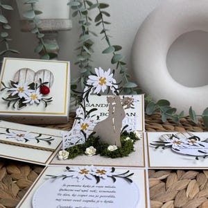 Op de afbeelding: Een witte en gouden trouwkaart met een pop-up ontwerp met een stel dat onder een bloemenboog staat. De kaart heeft een witte achtergrond met gouden accenten en een bericht dat luidt "Se pomnita kako je vaju mladi, Gubezni val ujel tak, iznenadia, Ta dan bo zopet sta jo potrdila, maj veter wet zapiha ji v krila, Cestitke, mladoporočenca! Klemen in Spela Bodnik".