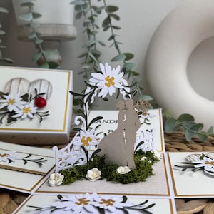 Op de afbeelding: Een witte en gouden trouwkaart met een pop-up ontwerp met een stel dat onder een bloemenboog staat. De kaart bevat een witte cirkel met een bericht in een vreemde taal.