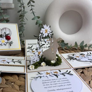 Op de afbeelding: Een witte en gouden trouwkaart met een pop-up ontwerp met een bruid en bruidegom onder een bloemenboog. De kaart heeft een witte achtergrond met gouden accenten en een boodschap in een cirkel die luidt "Se pomnita kak je vaju mlada, gubezni val ujel tak, iznenada Ta dan, ky copet sta jo potrdila, naj veter ovet zapika ji v krila. Costicks, mladoporočenca Klemen in Spela Botnik."