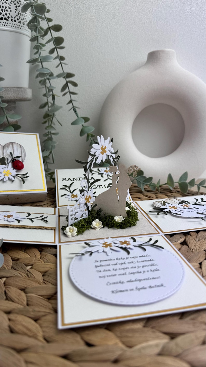 Op de afbeelding: Een witte en gouden trouwkaart met een pop-up ontwerp met een bruid en bruidegom onder een bloemenboog. De kaart heeft een witte achtergrond met gouden accenten en een boodschap in een cirkel die luidt "Se pomnita kak je vaju mlada, gubezni val ujel tak, iznenada Ta dan, ky copet sta jo potrdila, naj veter ovet zapika ji v krila. Costicks, mladoporočenca Klemen in Spela Botnik."
